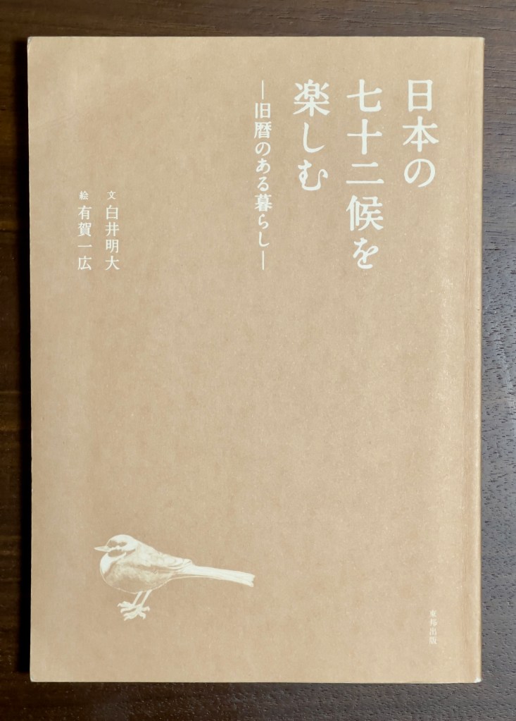 Tan cover of a book with (all in white print) a bird in the bottom left corner. The writing is in Japanese, so read vertically right to left. 『日本の七十二候を楽しむー旧暦のある暮らしー』on the upper right and 『文白井明大』and 『有賀一広』on the left.