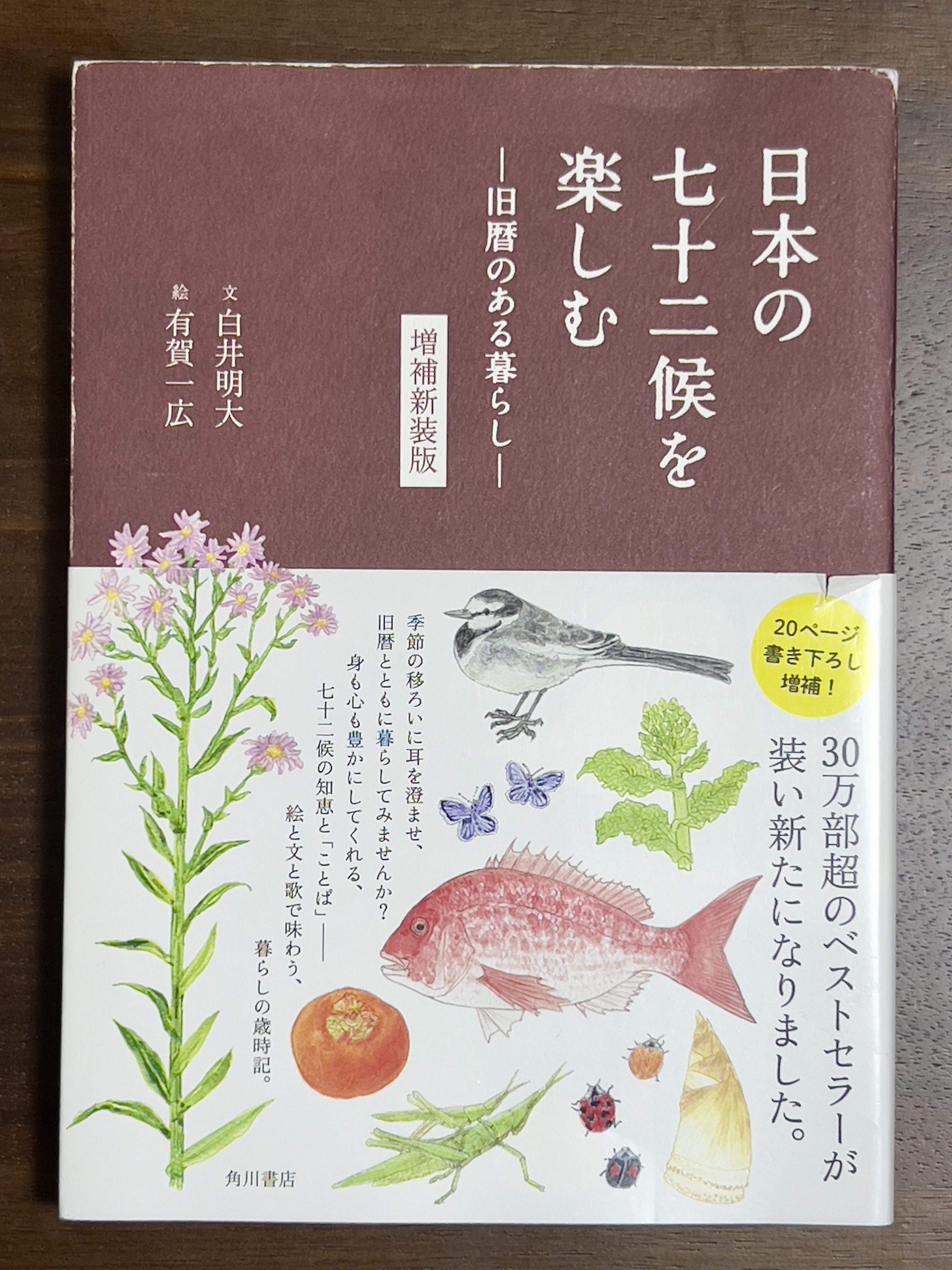 Dust jacket of Enjoying Japan's 72 Seasons - Living with the Lunar Calendar. Maroon and white, with colored illustrations of flowers/plants, fruit/vegetables, insects, a bird, and a fish. The title is in Japanese. 『日本の七十二候を楽しむー旧暦のある暮らしー』in the upper right and the author and illustrator in the upper left 『文白井明大』and 『有賀一広』. In the middle 『増補新装版』.