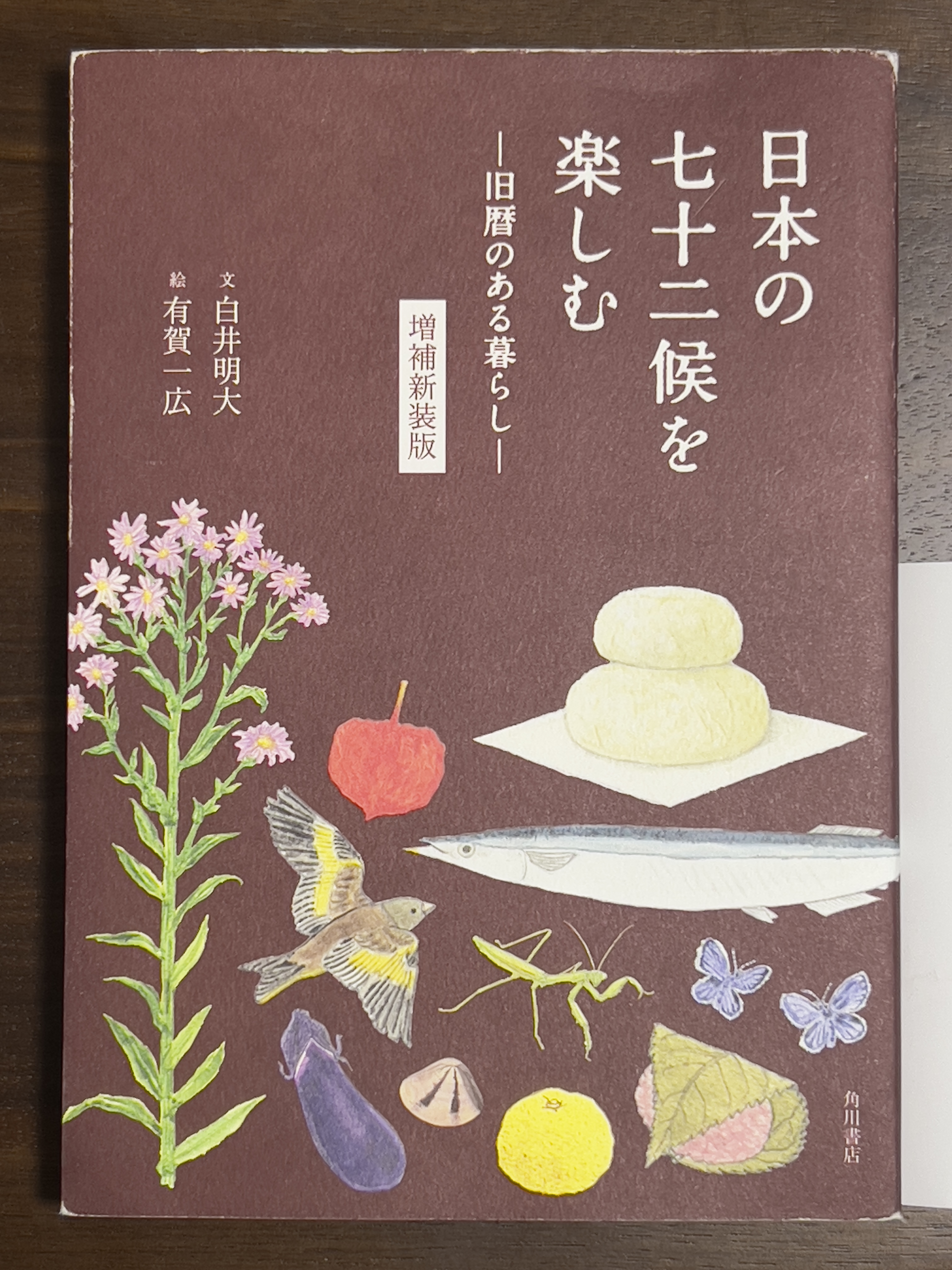 Dust jacket of Enjoying Japan's 72 Seasons - Living with the Lunar Calendar. Maroon with colored illustrations of flowers/plants, fruit/vegetables, Japanese sweets, insects, a bird, and shellfish. The title is in Japanese. 『日本の七十二候を楽しむー旧暦のある暮らしー』in the upper right and the author and illustrator in the upper left 『文白井明大』and 『有賀一広』. In the middle 『増補新装版』.